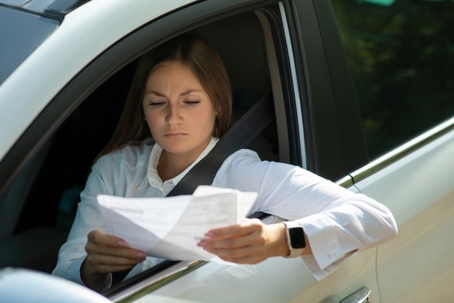 Driver reading paperwork in a car, representing license suspension and financial stress related to unpaid debt.